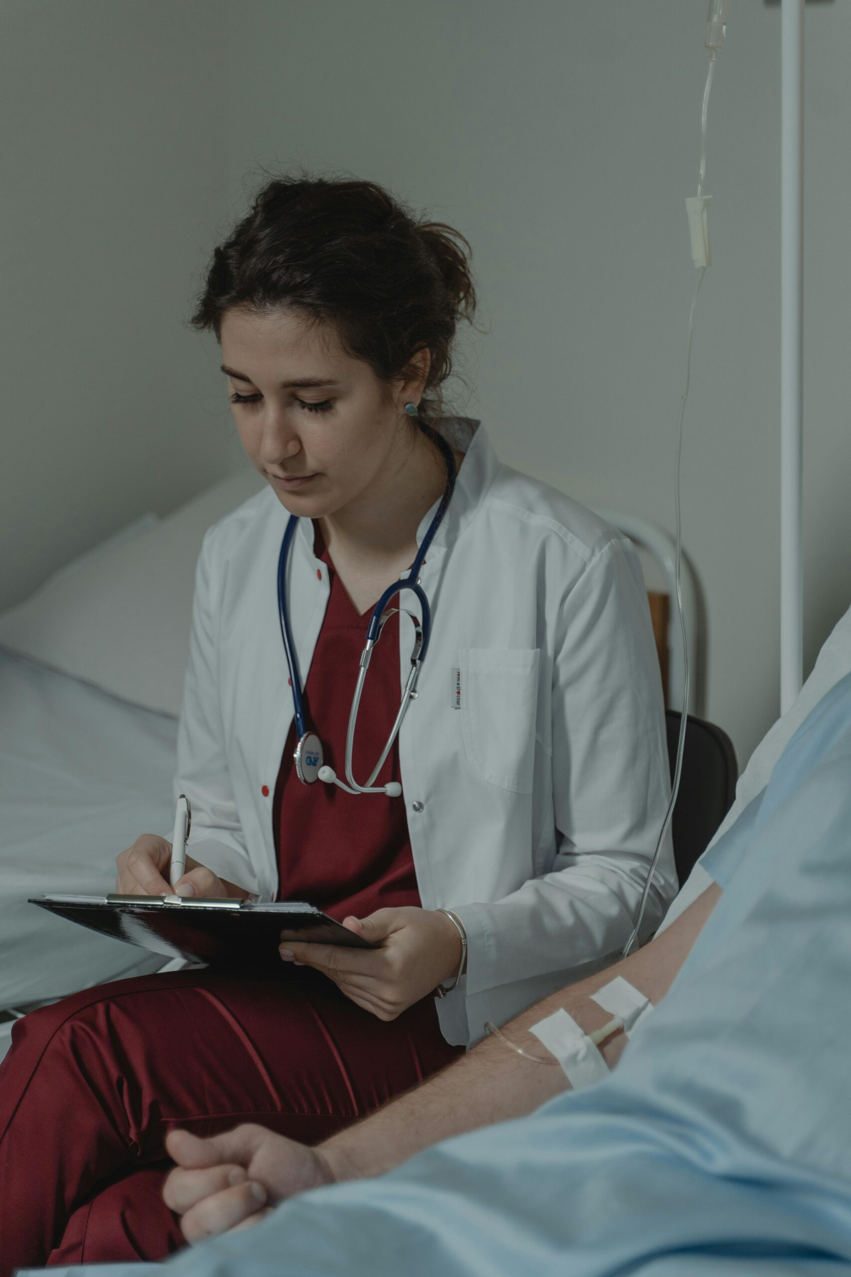 Medical professional with stethoscope taking notes beside patient in hospital setting.